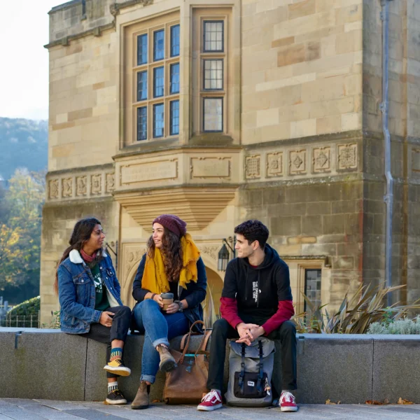 Three students sit in front of a historic building on Bangor University campus.
